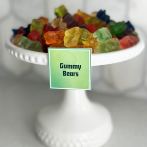 May include: A white cake stand with a variety of colorful gummy bears on top. A green label with the text "Gummy Bears" is attached to the stand.