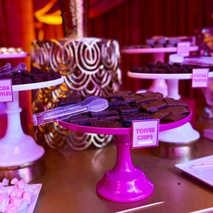 Puede incluir: Un soporte para pastel rosa con un plato blanco que contiene trozos de caramelo de chocolate. El soporte está sobre una mesa dorada con otros soportes para pastel que contienen trufas de chocolate y otras golosinas.