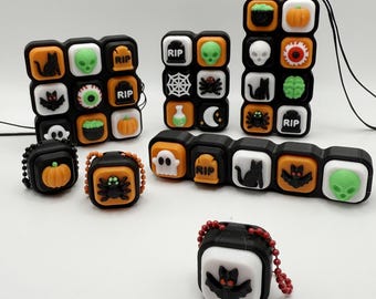 May include: A collection of Halloween-themed accessories, featuring black, white, and orange color schemes. The items include keychains and decorative blocks with spooky icons like pumpkins, bats, skulls, and tombstones. The accessories are designed with a playful, festive aesthetic.