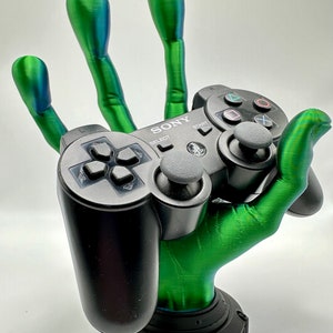 Könnte beinhalten: Eine grüne, im 3D-Druck hergestellte Hand hält einen schwarzen Sony Playstation 3-Controller. Die Hand hält den Controller in Spielposition. Der Controller trägt die Aufschriften "SONY", "SELECT" und "START".