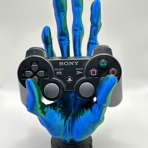 May include: A 3D printed hand, painted in a blue and green gradient, holding a black PlayStation 3 controller. The hand is posed with the fingers extended, as if holding the controller up.