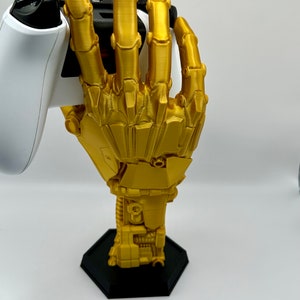 Op de afbeelding: Een gouden robot hand die een witte videogamecontroller vasthoudt. De hand is bevestigd aan een zwarte zeshoekige basis.