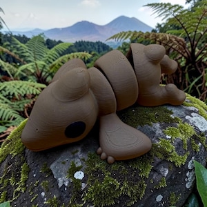 May include: A brown, articulated wooden sculpture of a creature with a segmented body, four limbs, and a large head with a black eye. The sculpture is resting on a moss-covered rock, with a backdrop of lush green ferns and mountains.
