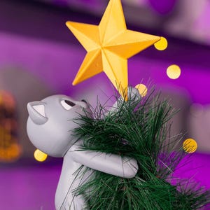 May include: A grey cat figurine holding a green Christmas tree with a yellow star on top. The cat is looking up at the star. The background is a blurred purple with yellow bokeh lights.