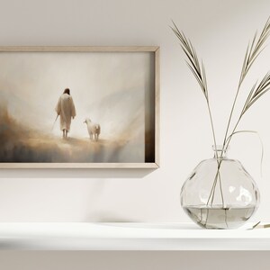 May include: A framed painting of a person in a white robe walking with a white lamb in a misty landscape. The painting is hanging on a white wall above a white shelf with a clear glass vase filled with dried grass.