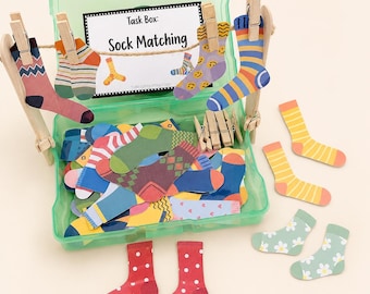 May include: A green plastic storage box containing a collection of colorful paper sock cutouts with various patterns. A wooden stand with twine and clothespins holds several socks, and a label reads Task Box: Sock Matching.