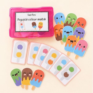 以下が含まれることがあります： Task Box: Popsicle colour matchとラベルが貼られた鮮やかなピンク色のプラスチック製収納ボックス。笑顔の顔が描かれたアイスキャンディー型の切り抜きと様々な色の組み合わせ、そしてアイスキャンディーの輪郭が描かれた長方形のカードが複数入っています。
