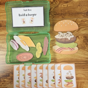 Könnte beinhalten: Eine grüne Plastikbox mit dem Text "Task Box: Build a burger" enthält Burgerzutaten. Ein fertiger Burger und Karten mit Burger-Illustrationen liegen auf einer Holzoberfläche. Zu den Zutaten gehören eine Tomatenscheibe, Käse und Fleisch. Die Karten sind für ein Spiel.