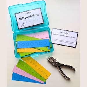 Peut inclure: Une boîte de rangement en plastique turquoise vif contenant des bandes de papier colorées avec des cercles imprimés, étiquetée Task Box: Hole punch strips. Inclut une carte d'instructions et une perforatrice en métal pour les cercles.