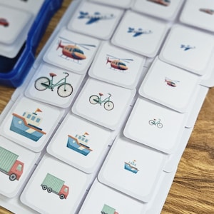 May include: A set of flashcards featuring various vehicles, including trains, airplanes, helicopters, bicycles, boats, and trucks. The cards are white with colorful illustrations and are labeled "Big Medium". A blue container is visible in the background.