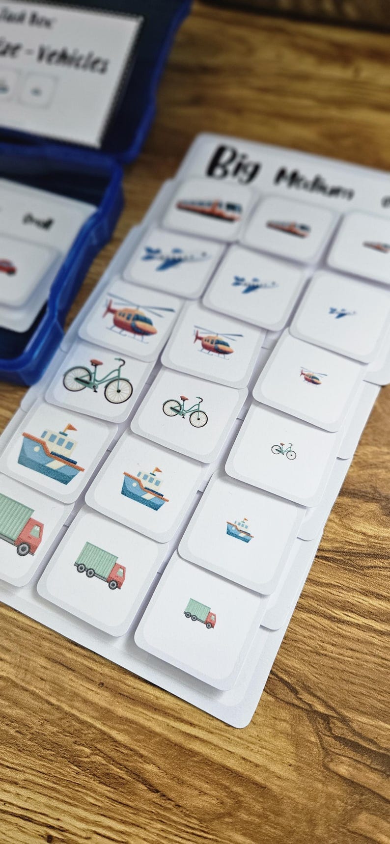 May include: A set of flashcards featuring various vehicles, including trains, airplanes, helicopters, bicycles, boats, and trucks. The cards are white with colorful illustrations and are labeled "Big Medium". A blue container is visible in the background.
