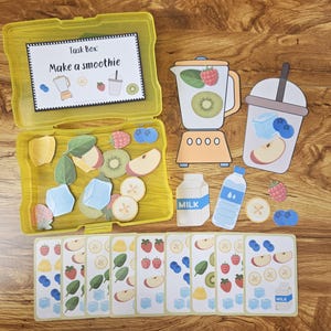 Może przedstawiać: Żółte pudełko z zadaniami z napisem "Make a smoothie" i różnymi wycinkami owoców, w tym kiwi, jabłko i truskawki. Zawiera również blender, kubek do smoothie, karton mleka, butelkę wody i karty ze składnikami smoothie.