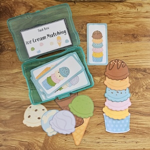 Może przedstawiać: Turkusowe plastikowe pudełko z kartami i wycięciami w kształcie lodów. Karty przedstawiają rożki lodów o różnych smakach. Tekst na pudełku brzmi "Task Box: Ice Cream Matching". Dodatkowe gałki i rożki lodów są rozrzucone.