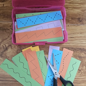 Könnte beinhalten: Eine rosa Plastikbox gefüllt mit bunten Papierstreifen mit gepunkteten und gestrichelten Linien. Eine Schere mit schwarzem Griff liegt auf der Holzoberfläche neben den Streifen, was auf eine Schneideaktivität hindeutet. Die Streifen sind in verschiedenen Farben.