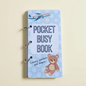 Op de afbeelding: Een rechthoekig pocket-activiteitenboek gebonden met drie zilveren metalen ringen. De kaft is lichtblauw met witte stippen, voorzien van een blauw vliegtuig, een bruine teddybeer en de tekst POCKET BUSY BOOK Colours, numbers and shapes.