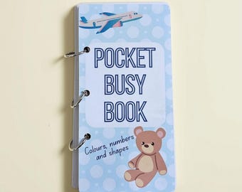 May include: A rectangular pocket busy book bound with three silver metal rings. The cover is light blue with white polka dots, featuring a blue airplane, a brown teddy bear, and the text POCKET BUSY BOOK Colours, numbers and shapes.