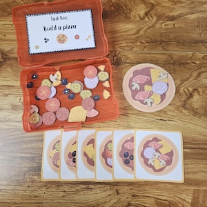 May include: An orange task box labeled "Build a pizza" with various pizza topping cutouts, including mushrooms, olives, and onions. Several pizza cards are arranged below the box, and a single pizza cutout is to the right.