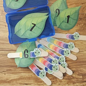 Könnte beinhalten: Lehrreiches Zählset mit grünen Blattausschnitten mit den Zahlen 1-9, einem blauen Kunststoffbehälter und bunten Eisstielen mit Raupenmotiven. Die Blätter und Raupen sind auf einer Holzoberfläche angeordnet, ideal für frühe Lernaktivitäten.