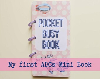 May include: A rectangular pocket busy book with a light pink background featuring white polka dots. The cover displays the text POCKET BUSY BOOK and My first ABCs Mini Book, with illustrations of a blue airship and a pink airplane, bound by three purple rings.
