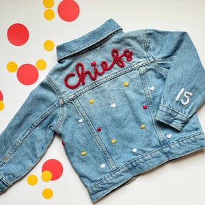 May include: A light blue denim jacket with the word "Chiefs" embroidered in red on the back. The jacket has the number "15" embroidered on the sleeve and is decorated with small heart and circle embellishments.