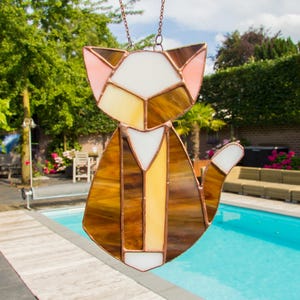 K&ouml;nnte beinhalten: Ein Buntglas-Katzen-Sonnenf&auml;nger mit braunen, wei&szlig;en und rosa Glasst&uuml;cken. Die Katze sitzt mit ihrem Schwanz um den K&ouml;rper gewickelt. Der Sonnenf&auml;nger h&auml;ngt an einer Kette.