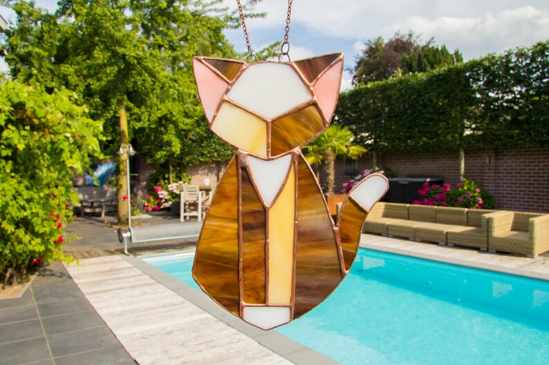 K&ouml;nnte beinhalten: Ein Buntglas-Katzen-Sonnenf&auml;nger mit braunen, wei&szlig;en und rosa Glasst&uuml;cken. Die Katze sitzt mit ihrem Schwanz um den K&ouml;rper gewickelt. Der Sonnenf&auml;nger h&auml;ngt an einer Kette.