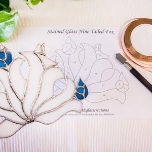 Op de afbeelding: Een wit en blauw gebrandschilderd negenstaartige vos, met een zilveren omtrek, vastgehouden door een hand. Een patroon, koperen tape en een glassnijder liggen op de tafel. De tekst op het patroon luidt "Stained Glass Nine Tailed Fox".