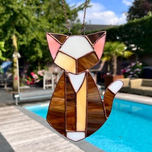 K&ouml;nnte beinhalten: Ein Buntglas-Sonnenf&auml;nger in Form einer Katze mit braunen, wei&szlig;en und rosa Glasst&uuml;cken. Die Katze sitzt mit ihrem Schwanz um den K&ouml;rper gewickelt. Der Sonnenf&auml;nger h&auml;ngt an einer Kette.