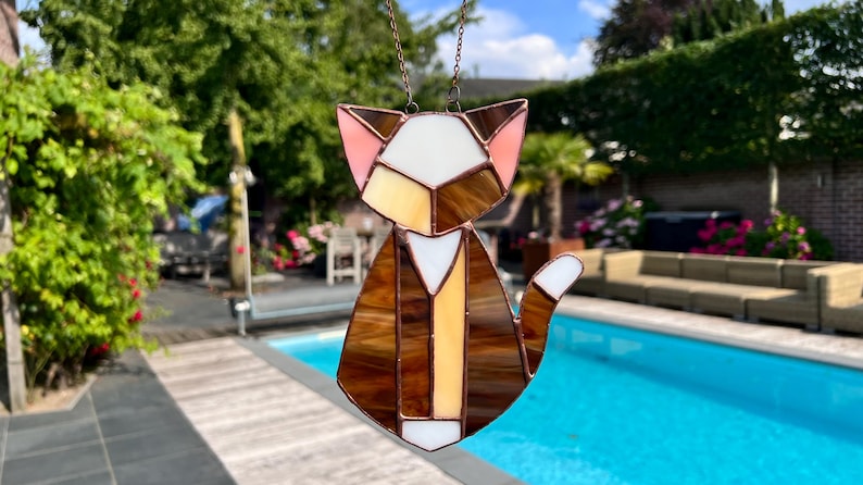 K&ouml;nnte beinhalten: Ein Buntglas-Sonnenf&auml;nger in Form einer Katze mit braunen, wei&szlig;en und rosa Glasst&uuml;cken. Die Katze sitzt mit ihrem Schwanz um den K&ouml;rper gewickelt. Der Sonnenf&auml;nger h&auml;ngt an einer Kette.