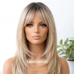 Puede incluir: Una peluca de longitud media con cabello rubio en capas, raíces más oscuras y flequillo desfilado que enmarca el rostro. El cabello tiene una textura lisa con reflejos sutiles. El texto LabelLaunch es visible en la parte central inferior.