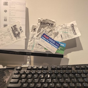 May include: A black plastic document holder clip attached to a computer keyboard, holding several white paper receipts from Costco and Super Save Foods, along with a blue and white FastSigns business card and a printed order summary sheet.