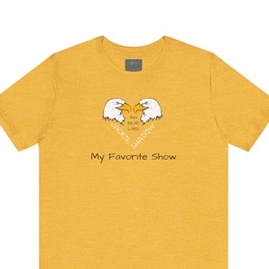 Könnte beinhalten: Ein gelbes T-Shirt mit einem Aufdruck von zwei sich gegenüberstehenden Weißkopfseeadlern. Die Adler bilden mit ihren Köpfen eine Herzform. Der Text "Big Bear Lake" befindet sich innerhalb der Herzform. Der Text "Jackie" befindet sich links neben dem Herzen und der Text "Shadow" rechts neben dem Herzen. Der Text "My Favorite Show" befindet sich unterhalb des Herz-Motivs.