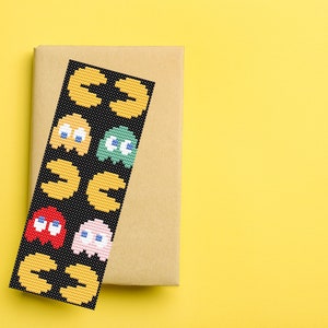 May include: A black bookmark with a repeating pattern of yellow Pac-Man characters and ghosts in various colors, including red, green, and pink. The bookmark is on a brown paper background.