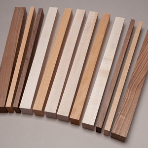 May include: A collection of 14 wooden dowels in various shades of brown, beige, and white. The dowels are arranged in a row from darkest to lightest.