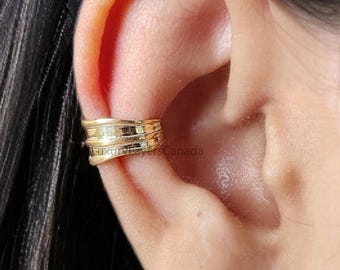Könnte beinhalten: Ein goldfarbener Ohrklemmen-Aufsatz mit einem geschichteten Design wird am Ohr getragen. Ein kleiner, quadratischer, klarer Ohrstecker ist ebenfalls sichtbar. Das Ohr ist vor einem Hintergrund aus dunklem Haar platziert.