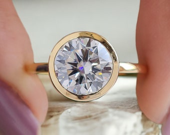 Op de afbeelding: Een gouden ring met een grote, ronde, bezel-gezet, heldere edelsteen. De ring ligt op een wit, getextureerd oppervlak.