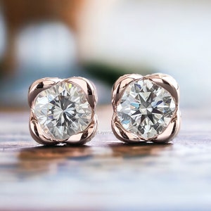 May include: A pair of rose gold stud earrings with a round, clear diamond in each setting. The diamonds are set in a square-shaped frame.