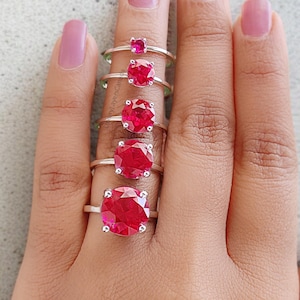 May include: Five silver rings with bright red gemstones are displayed on a hand. The rings, varying in size, have round, faceted gemstones. The largest ring is at the base of the finger, the smallest at the top. The gemstones reflect light.
