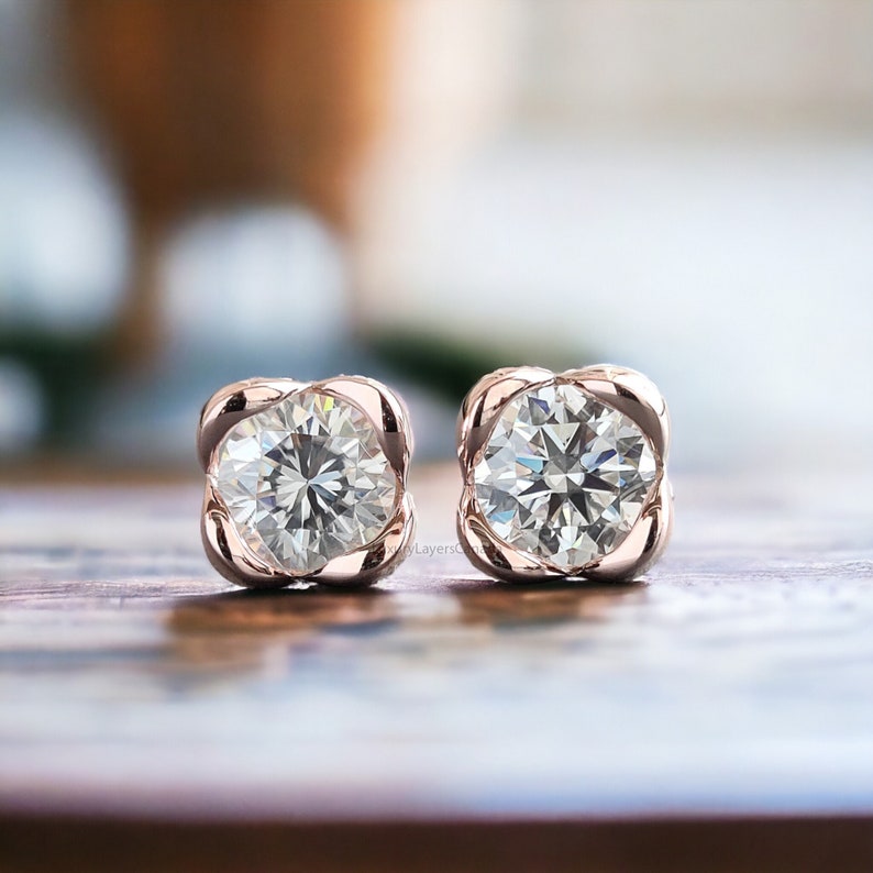 May include: A pair of rose gold stud earrings with a round, clear diamond in each setting. The diamonds are set in a square-shaped frame.