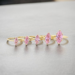 May include: Five gold rings with pink marquise-shaped gemstones. The rings are arranged in a row from smallest to largest.