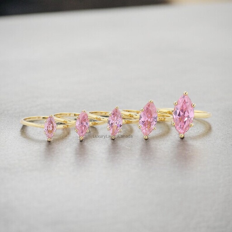 May include: Five gold rings with pink marquise-shaped gemstones. The rings are arranged in a row from smallest to largest.