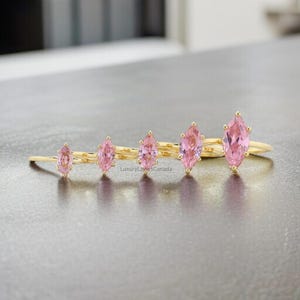 May include: Five gold rings with pink marquise-shaped gemstones. The rings are arranged in a row from smallest to largest.