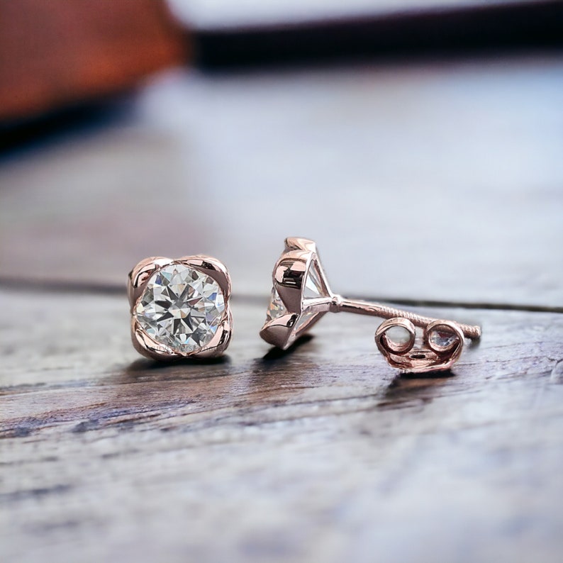 May include: A pair of rose gold stud earrings with a round, clear diamond in each setting. The earrings have a simple, elegant design.