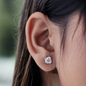 May include: A close-up of a person's ear with a small, round, diamond stud earring in a rose gold setting.