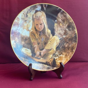 May include: A porcelain plate with a gold rim features a painting of a young girl with blonde hair sitting in a forest setting. The girl is wearing a yellow dress and has a blue ribbon in her hair. The background is a soft brown and green.