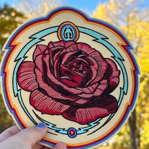 Peut inclure: Écusson brodé avec une grande rose rouge au centre, sur un fond crème. Le patch circulaire a une bordure décorative bleue, rouge et jaune. Un logo est visible en haut du patch.