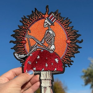 Puede incluir: Parche bordado con un esqueleto con sombrero de copa, sentado sobre una seta roja con lunares blancos. El esqueleto está sobre un diseño de sol naranja con llamas negras. El parche se sostiene contra un cielo azul.