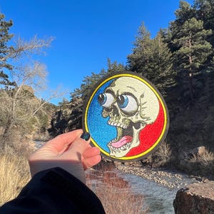 May include: A circular patch with a cartoon skull design. The skull has large, bulging eyes and an open mouth. The patch features blue and red sections with a yellow border. The skull is cream-coloured.