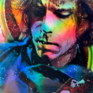 May include: A close-up, psychedelic image of a person's face, with rainbow-coloured light reflecting across the features. The person appears to be wearing a dark blue shirt. A guitar strap with skulls and roses is visible.