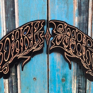 Könnte beinhalten: Zwei schwarze, flügelförmige Wanddekorationen mit aufwendigen goldfarbenen Schriftzügen und Designs. Die Dekorationen sind an einer verwitterten, türkis lackierten Holztür angebracht.